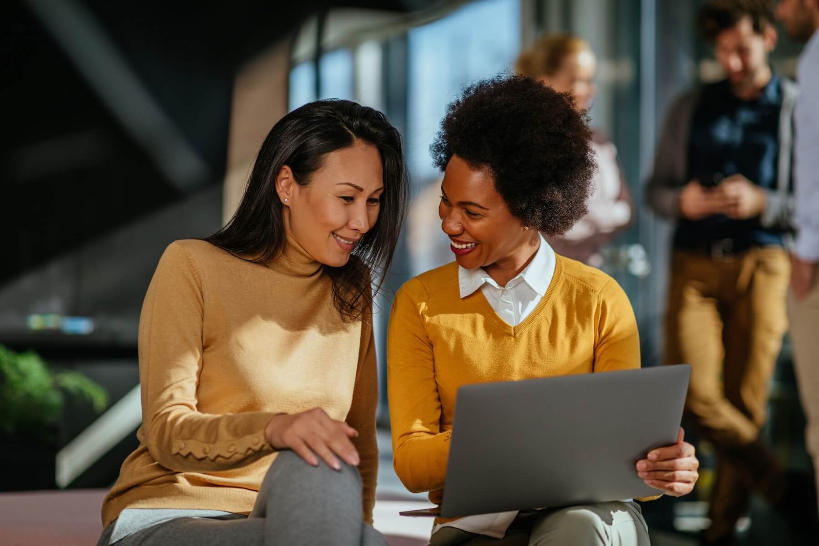 Two women smiling and engaging with a laptop, showcasing collaboration in a modern workspace, with a background of colleagues, emphasizing the importance of effective communication and customer support solutions.